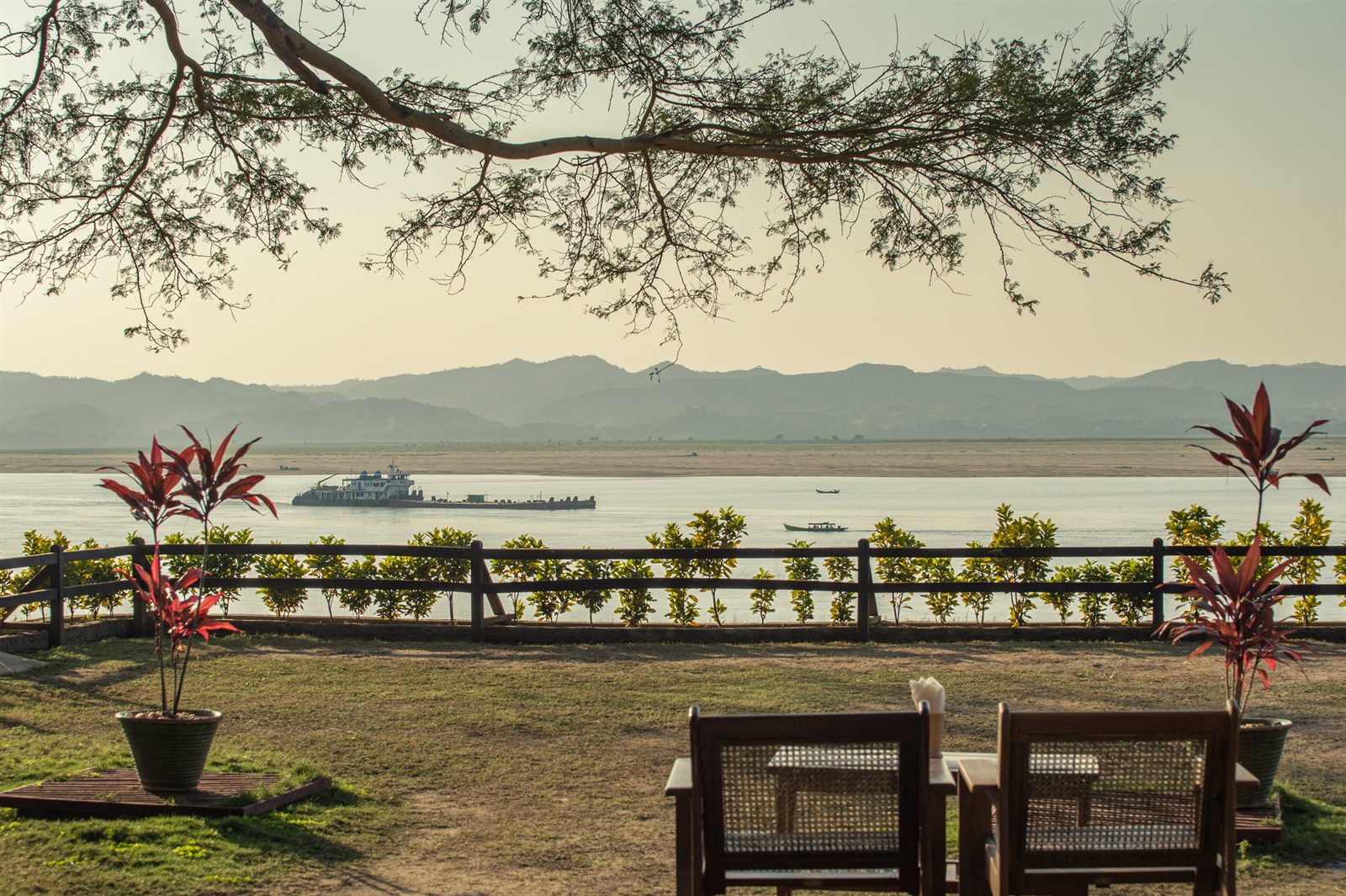 A riverboat on a wide river seen from a riverside garden with hills on the far bank, the kind of view an Irrawaddy cruise offers